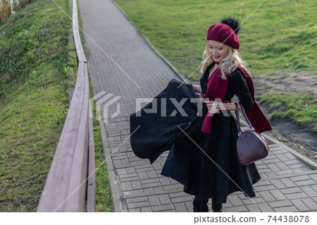 A cheerful girl walks up the stairs in a burgundy palette and biret, with a charismatic appearance in black clothes, in the fall against the background of trees there is a blue sky. 74438078