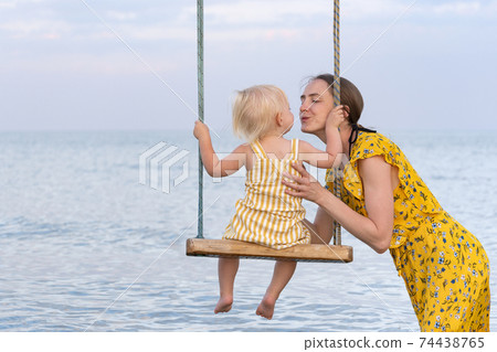 Young mother rolls baby on swing and kisses him. Tenderness and maternal care. Swing on the seashore. Young mother rolls baby on swing and kisses him. Tenderness and maternal care. Swing on the seashore. 74438765