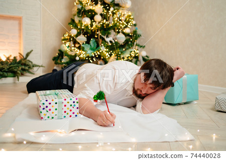 a man lies under the Christmas tree at home and fills out documents.  74440028