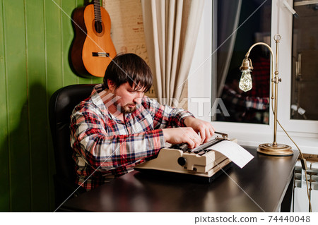 a man in plaid pajamas types on a typewriter at night in his home office. 74440048