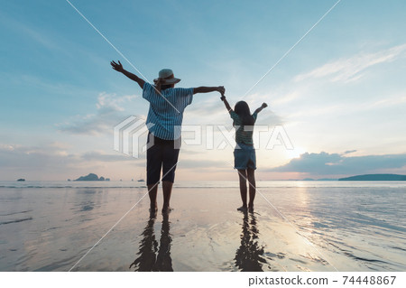 Silhouette of asian mother and daughter holding hand together standing on the beach looking at the beautiful sea and sky at the sunset time. 74448867