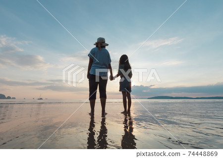Silhouette of asian mother and daughter holding hand together standing on the beach looking at the beautiful sea and sky at the sunset time. Silhouette of asian mother and daughter holding hand together standing on the beach looking at the beautiful sea and sky at the sunset time. 74448869