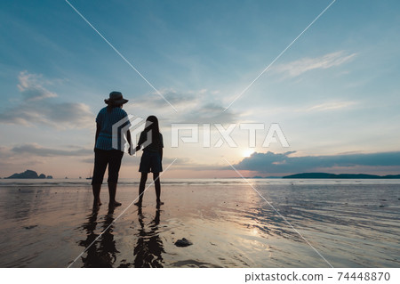 Silhouette of asian mother and daughter holding hand together standing on the beach looking at the beautiful sea and sky at the sunset time. 74448870