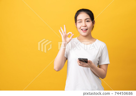 Cheerful beautiful Asian woman holding smartphone and shows ok sign on light yellow background. 74450686