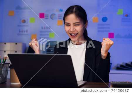 Happy excited Asian business woman celebrating success and working on laptop on the table at night. 74450687