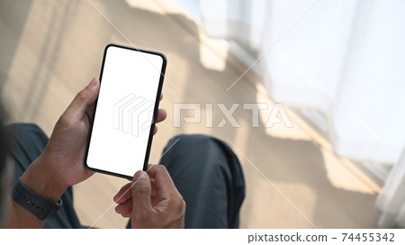 Close up view of young man holding mock up mobile phone with blank screen while sitting near window at home. 74455342