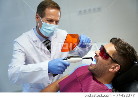 Focused dental specialist treating man with laser Focused dental specialist treating man with laser 74458377