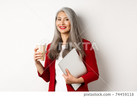 Business. Successful asian businesswoman with grey hair, wearing red blazer, drinking coffee and standing with laptop in hand, white background Business. Successful asian businesswoman with grey hair, wearing red blazer, drinking coffee and standing with laptop in hand, white background 74459118