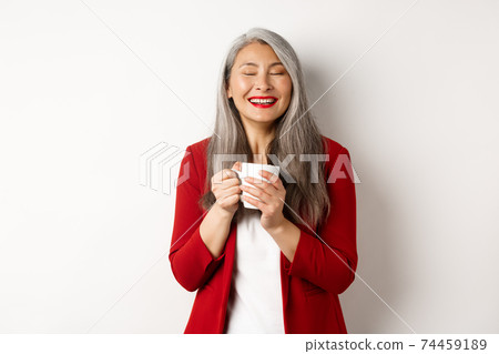 Business people concept. Happy office lady in red blazer enjoying drinking coffee, holding mug and smiling delighted, standing over white background Business people concept. Happy office lady in red blazer enjoying drinking coffee, holding mug and smiling delighted, standing over white background 74459189