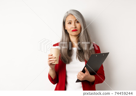 Tired and disappointed asian businesswoman with grey hair, drinking coffee in paper cup and looking gloomy at camera, standing over white background 74459244
