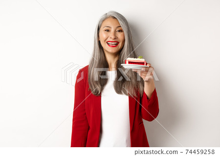 Celebration and holidays concept. Smiling asian businesswoman congratulate employees, giving plate with sweet cake and looking happy, standing over white background 74459258