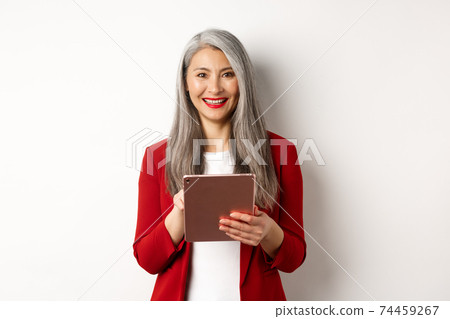 Business. Senior female entrepreneur working on digital tablet and smiling happy at camera, wearing red blazer and makeup, standing over white background 74459267