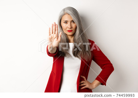 Serious asian woman in red blazer showing stop sign, telling no, frowning and rejecting something bad, standing against white background 74459993