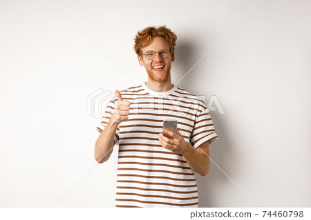 Technology and e-commerce concept. Satisfied male model with red hair, showing thumbs-up and holding smartphone, white background Technology and e-commerce concept. Satisfied male model with red hair, showing thumbs-up and holding smartphone, white background 74460798