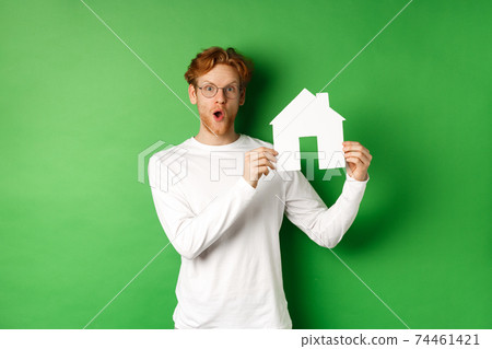 Real estate and buying property concept. Surprised young redhead man showing paper house model and looking amazed, standing over green background 74461421