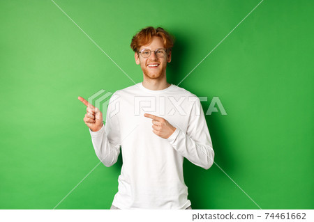 Handsome young man with red hair and beard smiling, pointing fingers left at logo, showing advertisement, standing over green background Handsome young man with red hair and beard smiling, pointing fingers left at logo, showing advertisement, standing over green background 74461662