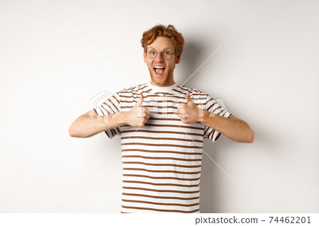 Young amazed man in red hair checking out something awesome, saying yes and showing thumbs-up, standing over white background 74462201