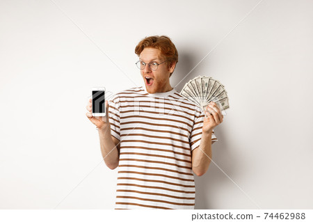 Amazed redhead man showing smartphone app on blank screen and money, winning prize cash online, standing over white background 74462988