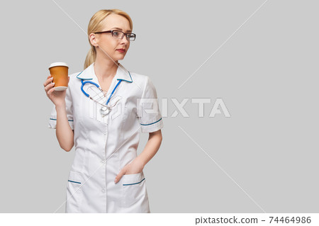 Portrait of a female doctor holding paper cup of coffee standing over grey background 74464986