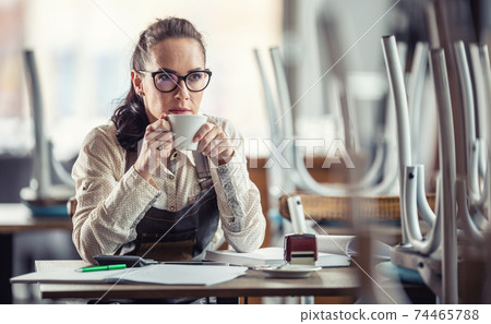 Business owner sips coffee sitting over accounting of her closed bar or restaurant Business owner sips coffee sitting over accounting of her closed bar or restaurant 74465788