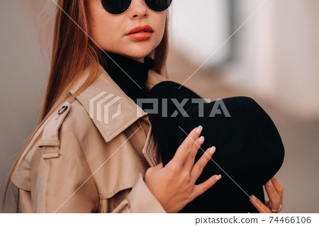 Stylish young woman in a beige coat and a black hat in his hands and glasses on a city street. Women's street fashion. Autumn clothing.Urban style. Stylish young woman in a beige coat and a black hat in his hands and glasses on a city street. Women's street fashion. Autumn clothing.Urban style. 74466106