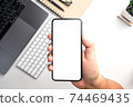 Top view of people hand holding smartphone with white blank screen over the  white table with smartphone, laptop,keyboard,notebook,pen,small tree and glasses.Flat lay design. 74469435