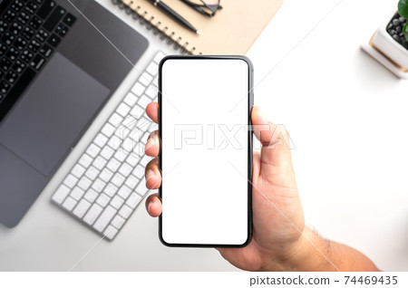 Top view of people hand holding smartphone with white blank screen over the  white table with smartphone, laptop,keyboard,notebook,pen,small tree and glasses.Flat lay design. 74469435