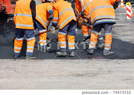 A team of road workers in orange reflective uniforms use shovels to level fresh asphalt on a clear day. A team of road workers in orange reflective uniforms use shovels to level fresh asphalt on a clear day. 74473510