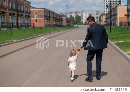 Rear view of a man in a black suit walks with his little daughter outdoors. A little girl in a white dress holds her father's hand. Paternal love Rear view of a man in a black suit walks with his little daughter outdoors. A little girl in a white dress holds her father's hand. Paternal love 74483559
