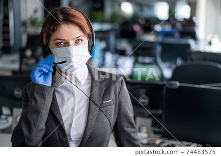 Female call center operator talking to a customer. Woman in mask and gloves at the workplace in the office. Social distance and safety measures to prevent infection. 74483575