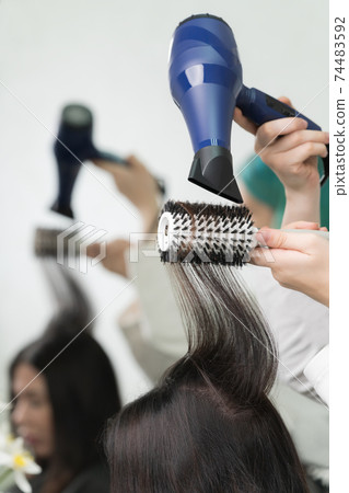 Hands of beautician dries long brunette hair of client using blue hair dryer and hairbrush in beauty salon Hands of beautician dries long brunette hair of client using blue hair dryer and hairbrush in beauty salon 74483592