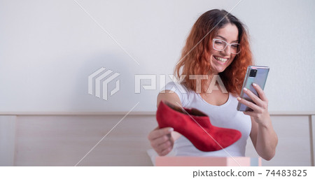 Online shopping using the application on the phone. Happy woman unpacks shoes and holds a smartphone. The expectation and reality of the purchase is consistent Online shopping using the application on the phone. Happy woman unpacks shoes and holds a smartphone. The expectation and reality of the purchase is consistent 74483825
