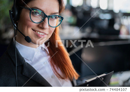 A friendly female support employee with glasses and a business suit is talking to a client on the headset. A woman with a folder in her hands answers the call with a smile. Happy worker Corporation. A friendly female support employee with glasses and a business suit is talking to a client on the headset. A woman with a folder in her hands answers the call with a smile. Happy worker Corporation. 74484107
