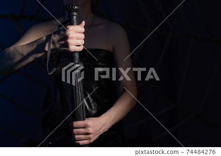BDSM concept. A woman in a leather corset holds a black lash with a long fringe. Role-playing games for adults. Close-up of sex toys for domination. The whip in female hands. 74484276