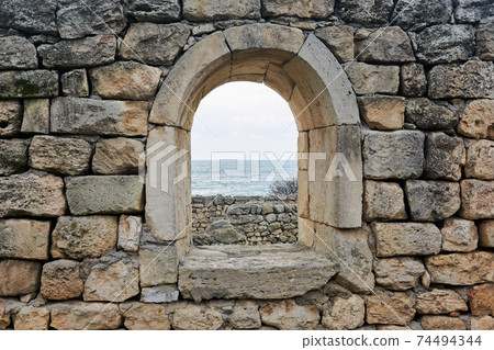 window opening in the ruins of an antique wall, behind which you can see the sea 74494344