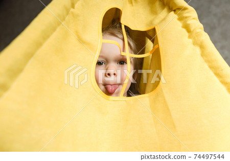 A little girl looks out the window of a teepee shows her tongue playing at home on the couch. A little girl looks out the window of a teepee shows her tongue playing at home on the couch. 74497544