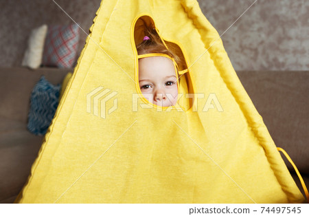 A little girl looks out the window of a teepee, plays at home on the couch. A little girl looks out the window of a teepee, plays at home on the couch. 74497545