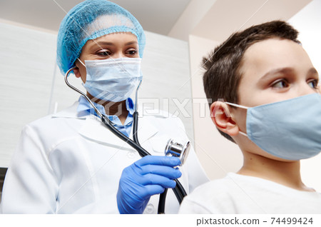 Female pediatrician in protective medical mask and gloves holding a stethoscope and auscultating a little patient. 74499424