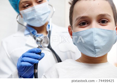 Smiling boy in medical mask during a pediatrician's examination. Pediatrist with stethoscope in hand ready to auscultate 74499425
