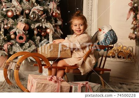 cute little blond baby girl in ivory dress on sledge on Christmas tree and fire place background cute little blond baby girl in ivory dress on sledge on Christmas tree and fire place background 74500228