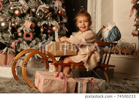 cute little blond baby girl in ivory dress on sledge on Christmas tree and fire place background cute little blond baby girl in ivory dress on sledge on Christmas tree and fire place background 74500233