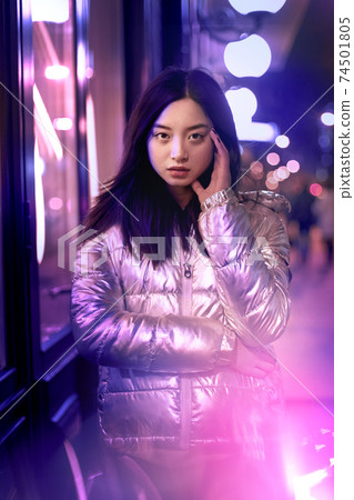 Neon Style. Asian girl in glossy jacket standing on the street in fluorescent violet lights touchign head concerned Neon Style. Asian girl in glossy jacket standing on the street in fluorescent violet lights touchign head concerned 74501805