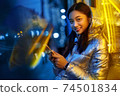 Neon Style. Asian girl in glossy jacket with wireless earphones standing on the street in fluorescent yellow lights listening to music browsing smartphone smiling cheerful 74501834