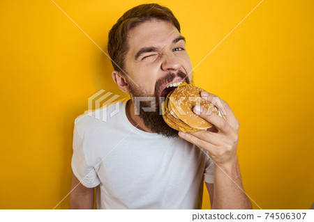 Man with hamburger in the hands of fast food diet food white t-shirt close-up yellow background Man with hamburger in the hands of fast food diet food white t-shirt close-up yellow background 74506307
