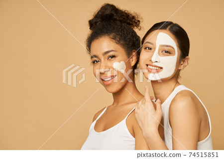 Two young female friends having fun, smiling at camera while posing with facial masks on isolated over beige background Two young female friends having fun, smiling at camera while posing with facial masks on isolated over beige background 74515851
