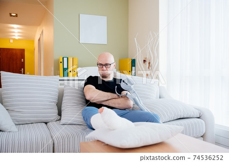 Angry man sitting on the sofa at home after the accident. He is holding crutches in his hand. Angry man sitting on the sofa at home after the accident. He is holding crutches in his hand. 74536252
