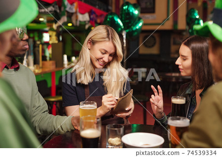Happy young blond waitress in workwear writing down orders of clients in notepad 74539334