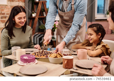 Cheerful young woman and little girl looking at man cutting sweet homemade pie Cheerful young woman and little girl looking at man cutting sweet homemade pie 74539461