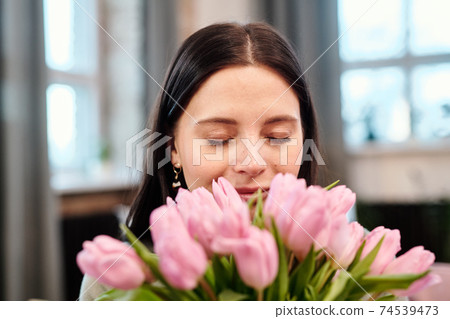 Happy young brunette female keeping her eyes closed while smelling pink tulips Happy young brunette female keeping her eyes closed while smelling pink tulips 74539473