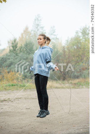 Blond active young woman in sportswear exercising with skipping-rope on path Blond active young woman in sportswear exercising with skipping-rope on path 74540382